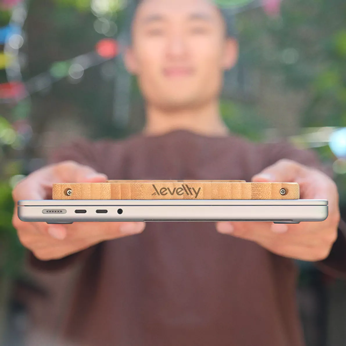 A hand holding a closed silver laptop with the folded bamboo Levelty stand resting on top